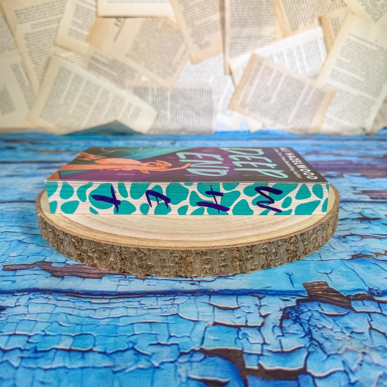 May include: A hardcover book titled "Deep End" by Ali Hazelwood, with a teal and white patterned edge, rests on a wooden slice. The book's cover features a woman with purple and orange tones. The background includes scattered book pages and a blue, textured surface.