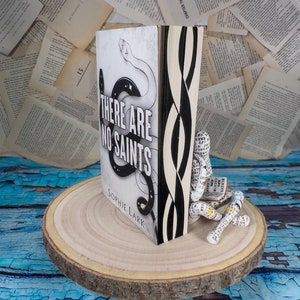 Può includere: Un libro tascabile in bianco e nero intitolato "There Are No Saints" di Sophie Lark. Il libro è in piedi su una fetta rotonda di legno. Una figura di cartapesta è seduta sulla fetta, leggendo un libro.
