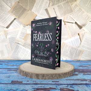 Può includere: Un libro con copertina rigida intitolato "FEARLESS" di Lauren Roberts. La copertina è nera con una corona argentata e accenti floreali rosa. Il libro è esposto su una fetta di legno con uno sfondo di pagine di libri sparse.