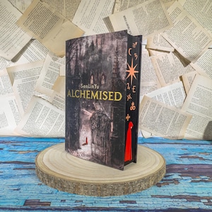 May include: A hardcover book titled "Alchemised" by SenLinYu. The cover features a dark, gothic illustration of a building with a gate and a figure in red. The spine has gold lettering and symbols, with a red bookmark.