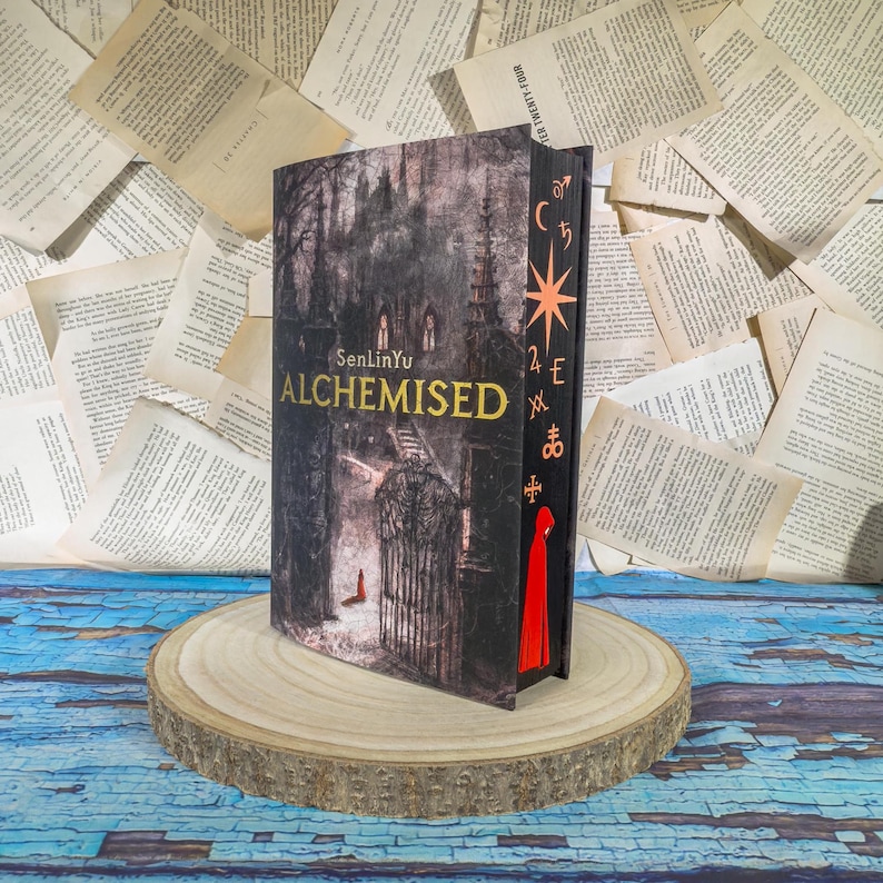 May include: A hardcover book titled "Alchemised" by SenLinYu. The cover features a dark, gothic illustration of a building with a gate and a figure in red. The spine has gold lettering and symbols, with a red bookmark.
