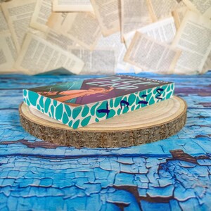 May include: A hardcover book with a teal and white patterned edge, resting on a wooden slice. The book cover features a woman with purple and orange tones. The book is on a blue, textured surface with scattered book pages in the background.