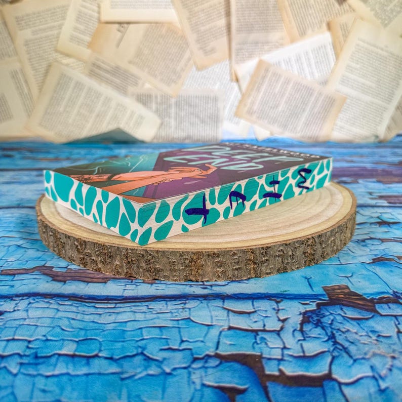 May include: A hardcover book with a teal and white patterned edge, resting on a wooden slice. The book cover features a woman with purple and orange tones. The book is on a blue, textured surface with scattered book pages in the background.