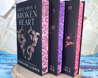 May include: A stack of hardcover books with black covers and decorative floral designs. The first book reads "ONCE UPON A BROKEN HEART" by Stephanie Garber. The spines feature pink, purple, and red patterns. The books are displayed on a wooden slice.
