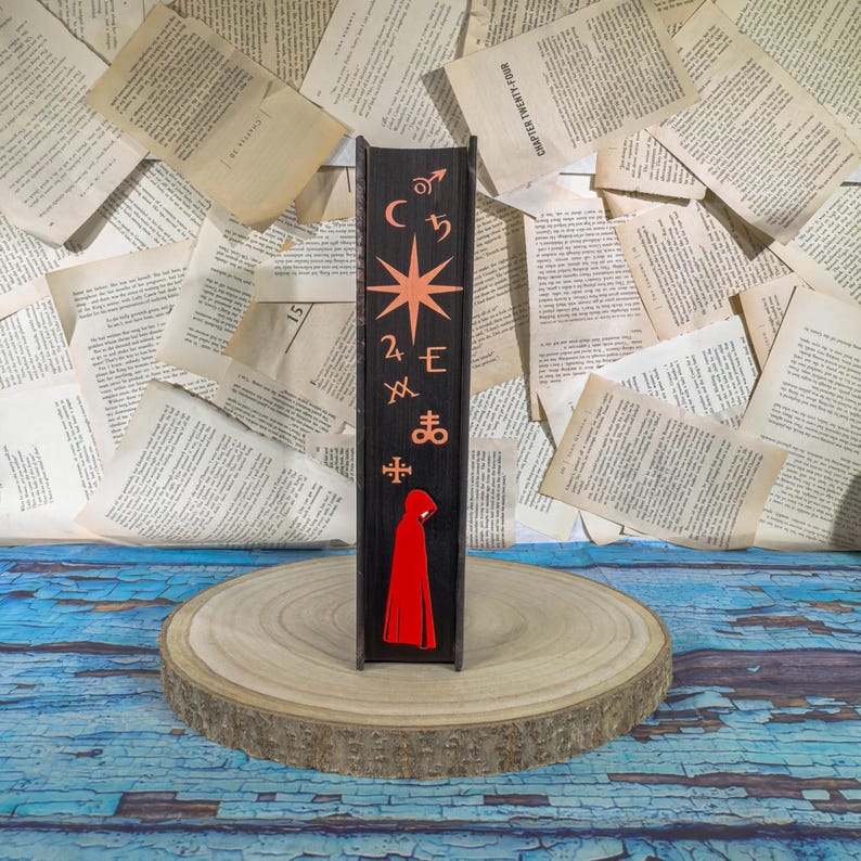 May include: A black wooden bookend with orange symbols and a star design. The bottom features a red hooded figure. The bookend is displayed on a wooden slice with a background of open book pages.