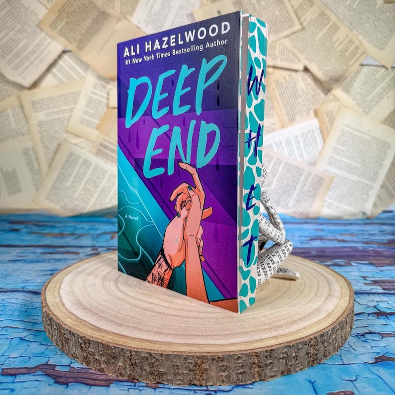 May include: A hardcover book titled "DEEP END" by Ali Hazelwood. The cover features a purple and teal design with two hands reaching for each other. The book is propped up on a wooden slice.