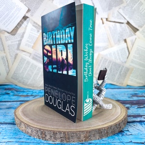 May include: A book titled "Birthday Girl" by New York Times bestselling author Penelope Douglas. The book cover features a woman in a blue swimsuit swimming in a pool. The book is propped up by a bookend shaped like a person reading a book.