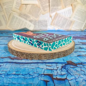 May include: A hardcover book titled "Deep End" by Ali Hazelwood, resting on a wooden slice. The book has a teal and white patterned edge and a colorful cover. The background features scattered book pages and a blue, textured surface.