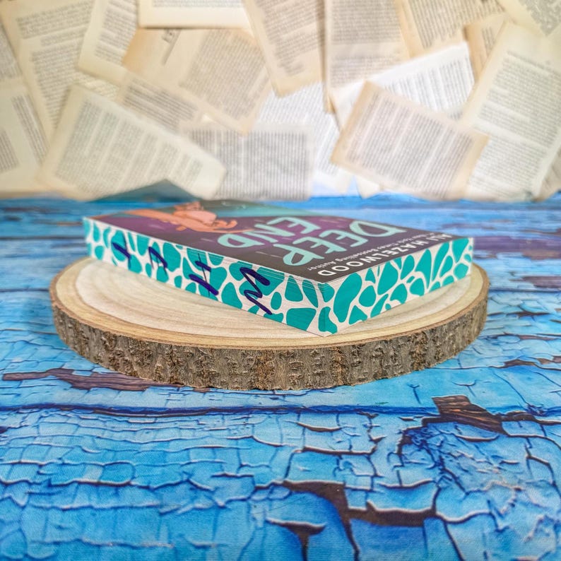 May include: A hardcover book titled "Deep End" by Ali Hazelwood, resting on a wooden slice. The book has a teal and white patterned edge and a colorful cover. The background features scattered book pages and a blue, textured surface.