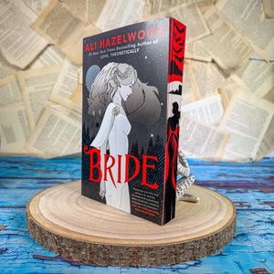 Może przedstawiać: Twarda oprawa książki zatytułowanej "Bride" autorstwa Ali Hazelwood. Okładka przedstawia kobietę w białej sukni z ilustracją wilka i księżyca. Grzbiet jest czerwony z czarnymi sylwetkami. Książka spoczywa na drewnianym plastrze.