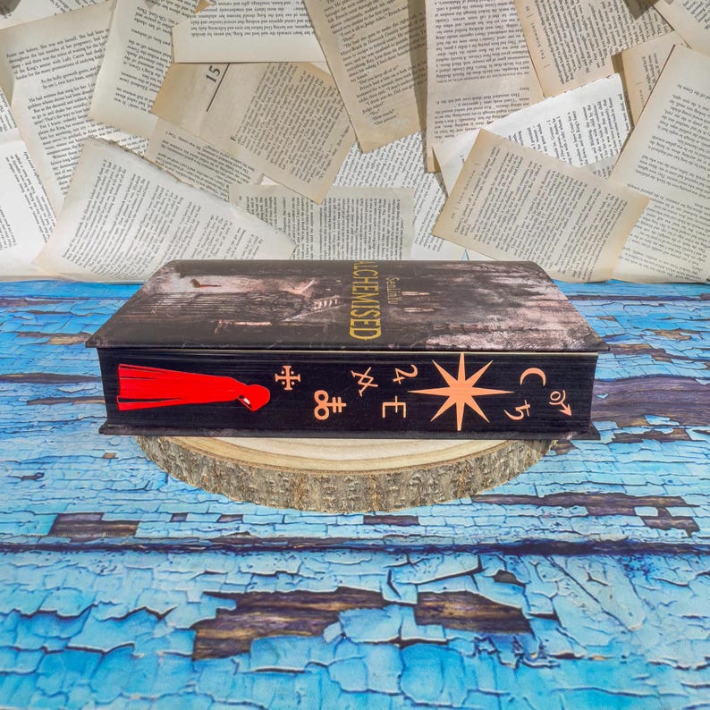 May include: A black wooden box with a distressed finish, designed to resemble a vintage book. The box features a red tassel and various celestial symbols. The top of the box is printed with the word "ALCHEMISED".