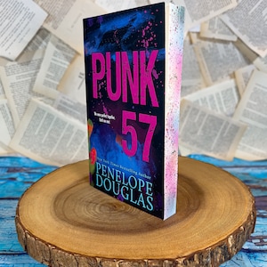 Op de afbeelding: Een paperback boek getiteld "Punk 57" van de New York Times bestseller auteur Penelope Douglas. De omslag heeft een zwarte achtergrond met roze en blauwe verfspatten en de titel in grote roze letters.
