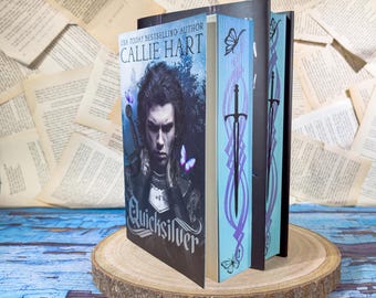 Könnte beinhalten: Ein gebundenes Buch mit dem Titel Quicksilver von Callie Hart, das ein düsteres Fantasy-Cover mit einer Person in Rüstung zeigt, sowie hellblaue Buchseitenkanten, die mit einem schwarzen Schwert, violetten Linien und Schmetterlingen verziert sind.
