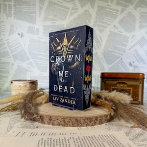 May include: A hardcover book titled Crown Me Dead by Liv Zander with a dark cover featuring a gold crown illustration, a skull motif, and white text. The book spine displays a red rose and gold crown icons against a black background.