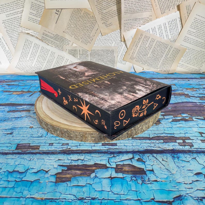 May include: A black book-shaped box with the title "Alchemised" in gold lettering. The box features orange decorative symbols and a starburst design. It rests on a wooden slice against a backdrop of scattered book pages and a blue, distressed wooden surface.