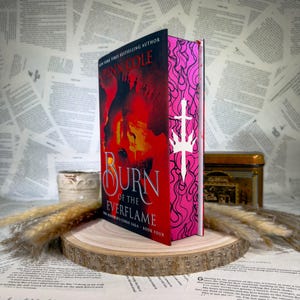 May include: A hardback book titled Burn of the Everflame by Penn Cole, featuring a red and black cover with a silhouette and a pink spine decorated with a white sword icon. The book is displayed upright on a circular wooden slice base.