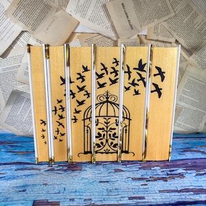 May include: A set of four books with gold-colored spines arranged side-by-side. The spines feature a continuous black graphic design of a birdcage in the center with black silhouettes of birds flying outward across the gold surfaces.