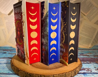 Könnte beinhalten: Vier Taschenbücher mit einem Halbmond-Design auf den Rückseiten. Die Bücher sind auf einer runden Holzscheibe gestapelt. Die Bücher sind betitelt mit "Crescent City", "House of Earth and Blood", "A Court of Thorns and Roses" und "A Court of Mist and Fury".