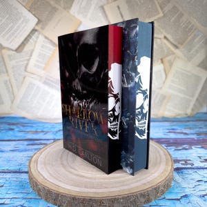 Könnte beinhalten: Zwei Hardcover-Bücher mit schwarzen Einbänden und einem Totenkopf-Design. Das Buch links trägt den Titel "Shallow River" und den Namen des Autors "H.D. Carlton". Die Bücher liegen auf einer runden Holzscheibe.
