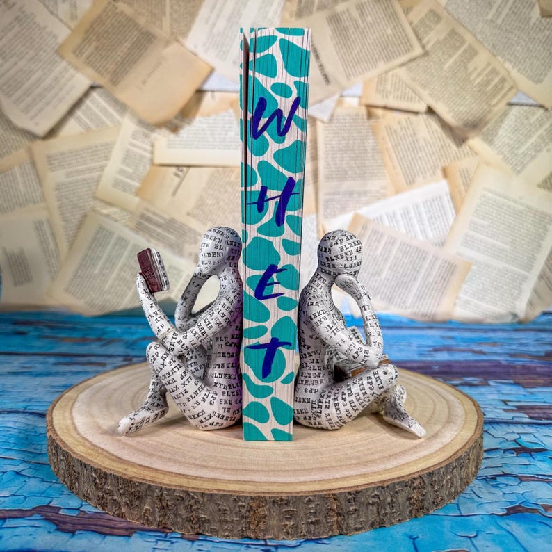 May include: A decorative bookend set featuring two figures made of paper with text, flanking a book with the word "Whet" in teal. The bookend set is on a wooden slice, with a background of open book pages.