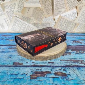 May include: A black book-shaped box with the title "ALCHEMISED" in gold lettering. The box features a red tassel and decorative symbols. It rests on a wooden slice against a backdrop of book pages and a blue, distressed surface.