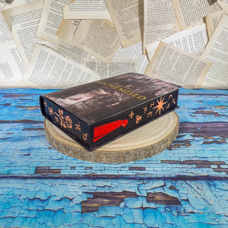 May include: A black book-shaped box with the title "ALCHEMISED" in gold lettering. The box features a red tassel and decorative symbols. It rests on a wooden slice against a backdrop of book pages and a blue, distressed surface.