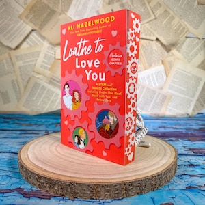 May include: A red book titled "Loathe to Love You" by Ali Hazelwood, featuring illustrations within red gear-shaped frames. The book is displayed on a wooden slice against a backdrop of open book pages.