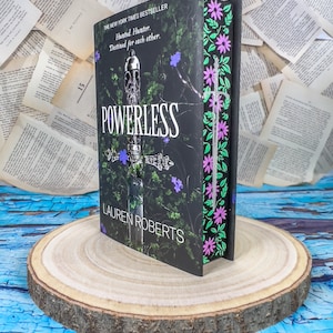 Può includere: Un libro con copertina rigida intitolato "POWERLESS" di Lauren Roberts, con una copertina nera con un disegno di spada e fiori viola. Il libro è esposto su una fetta di legno, con pagine sullo sfondo.