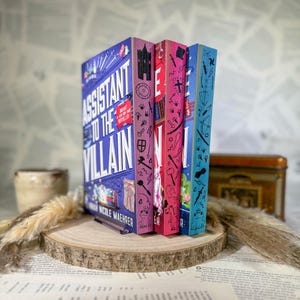 Könnte beinhalten: Drei gebundene Bücher stehen auf einer Holzscheibe. Das erste Buch ist blau mit dem Titel Assistant to the Villain von Hannah Nicole Maehrer. Das zweite Buch ist rosa und das dritte hellblau, beide mit verschiedenen schwarzen Strichzeichnungen.