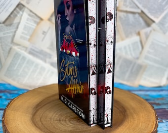 May include: A paperback book titled "Satan's Affair" by H.D. Carlton. The cover features a woman with dark hair and a red and white circus tent. The spine has a black and white design with a Ferris wheel and skulls.