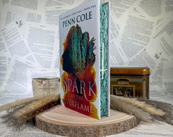 May include: A hardcover book titled Spark of the Everflame by Penn Cole, part of The Kindred's Curse Saga. The cover features a dark silhouette against a vibrant orange and teal abstract background. The book spine displays a white sword graphic.