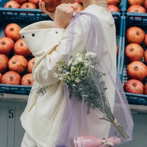 May include: A sheer lavender mesh tote bag containing a bouquet of white and yellow wildflowers and a small pink bottle of JP Chenet wine. The bag features a small black logo at the bottom with the text Holy Bags and a drawstring closure.