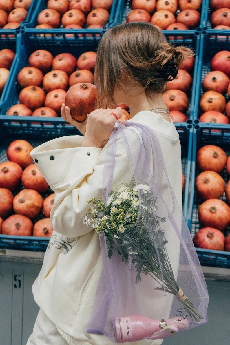 May include: A sheer lavender mesh tote bag containing a bouquet of white and yellow wildflowers and a small pink bottle of JP Chenet wine. The bag features a small black logo at the bottom with the text Holy Bags and a drawstring closure.