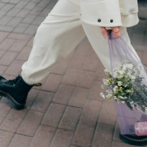 May include: A pair of cream-colored sweatpants with cuffed ankles, black leather lace-up boots with yellow stitching, and a sheer purple mesh tote bag containing a bouquet of white and yellow wildflowers and a small pink bottle.
