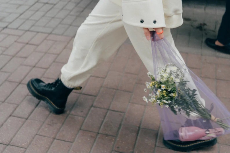 May include: A pair of cream-colored sweatpants with cuffed ankles, black leather lace-up boots with yellow stitching, and a sheer purple mesh tote bag containing a bouquet of white and yellow wildflowers and a small pink bottle.