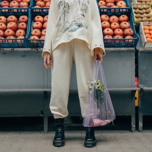 May include: A cream-colored sweatshirt with a graphic print and matching cream-colored sweatpants. A sheer, light purple mesh tote bag contains a small bouquet of white flowers and a pink bottle. Black lace-up boots complete the ensemble.