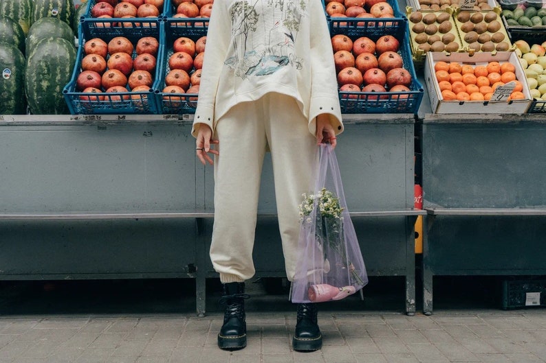 May include: A cream-colored sweatshirt with a graphic print and matching cream-colored sweatpants. A sheer, light purple mesh tote bag contains a small bouquet of white flowers and a pink bottle. Black lace-up boots complete the ensemble.