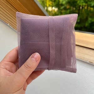 May include: A square-shaped sachet pouch made of mauve-colored fine mesh netting fabric. The small, lightweight bag features a matching mauve fabric ribbon band wrapped vertically around the center, securing the contents inside the semi-transparent material.