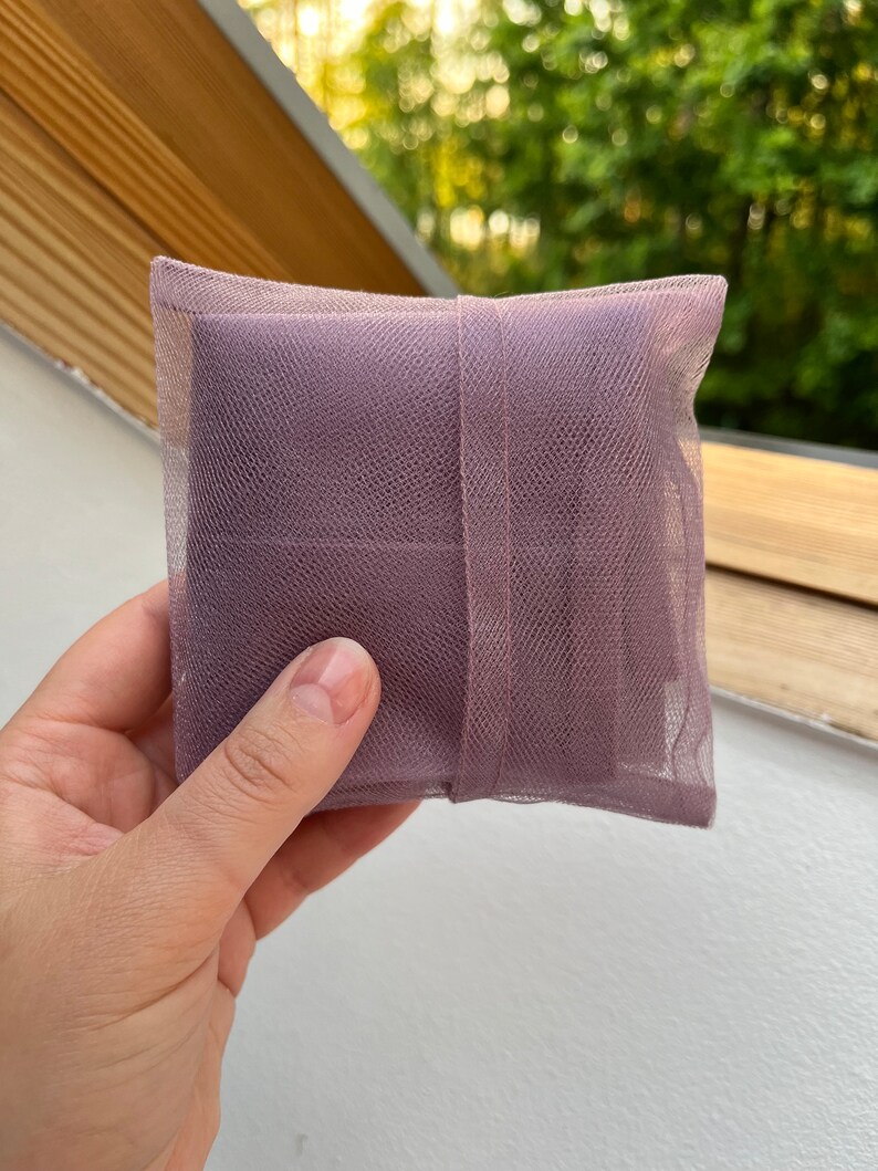 May include: A square-shaped sachet pouch made of mauve-colored fine mesh netting fabric. The small, lightweight bag features a matching mauve fabric ribbon band wrapped vertically around the center, securing the contents inside the semi-transparent material.
