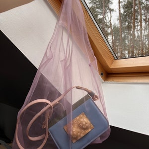 May include: A sheer lavender mesh tote bag containing a small rectangular blue crossbody bag with a light pink strap and a beige square patch featuring a white floral pattern. The mesh bag is made of a lightweight, semi-transparent fabric.
