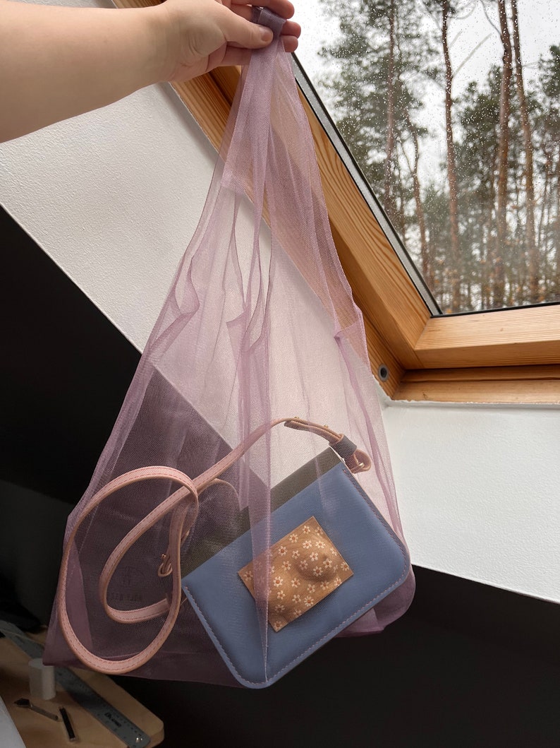 May include: A sheer lavender mesh tote bag containing a small rectangular blue crossbody bag with a light pink strap and a beige square patch featuring a white floral pattern. The mesh bag is made of a lightweight, semi-transparent fabric.