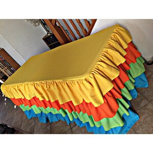 May include: A rectangular table covered with a yellow tablecloth and a tiered, ruffled skirt. The skirt features layers of yellow, orange, green, and blue fabric. The table is set indoors, with a glimpse of a staircase and other furniture in the background.