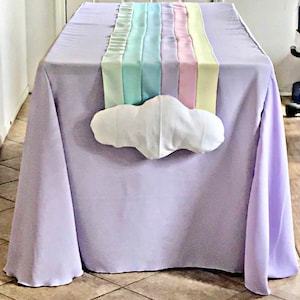 May include: A lavender tablecloth with a rainbow runner and a white cloud pillow. The rainbow runner is made of multiple pastel colored ribbons.