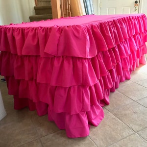 May include: A bright pink rectangular table cover with multiple layers of ruffled fabric. The table skirt is a vibrant fuchsia color, creating a bold and eye-catching design. The ruffles add texture and volume to the table covering.