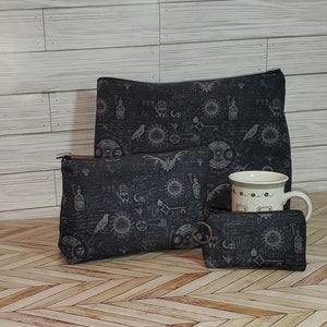 May include: Three black zippered pouches with a dark gray and white pattern of symbols, a bird, and a bat. The pouches are arranged in a pyramid shape with the largest pouch at the top. A white ceramic mug with a black and white pattern is in front of the pouches.
