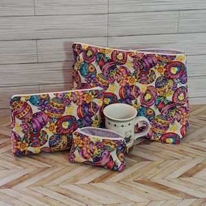 May include: Three zippered pouches with a colorful Easter egg pattern. The pouches are made of fabric and have a white background with purple, yellow, and pink Easter eggs. The largest pouch is standing upright, while the two smaller pouches are lying flat. A white mug with a brown bear design is sitting in front of the pouches.