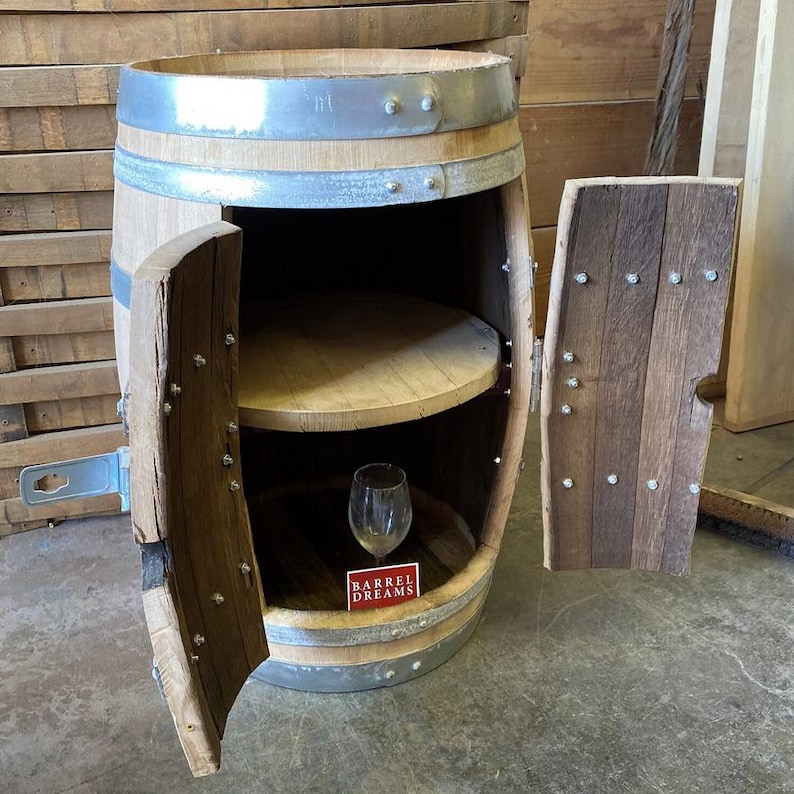 May include: A rustic, wooden barrel cabinet with a hinged door and a shelf inside. The cabinet is made from reclaimed wood and metal bands. A wine glass sits inside the cabinet. The text "BARREL DREAMS" is visible on a red label.