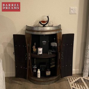 May include: A wooden barrel bar cabinet with two doors, a shelf, and a glass top. The cabinet is filled with various bottles of liquor, including Jack Daniels, Gentleman Jack, Jameson Black Barrel, and Knob Creek. The cabinet is decorated with a globe decanter and two glasses.