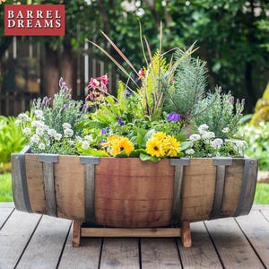 May include: A wooden barrel planter filled with vibrant flowers and greenery. The planter is made from reclaimed wood with metal bands and sits on a wooden stand. The flowers include yellow sunflowers, purple and white blooms, and various green foliage.