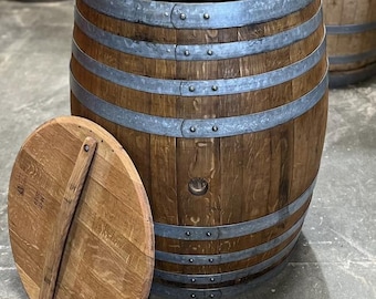 May include: A large, cylindrical brown wooden barrel with a hollow interior and an open top. The barrel is reinforced with six horizontal galvanised steel bands secured by rivets. A separate circular wooden lid with a central handle rests nearby.